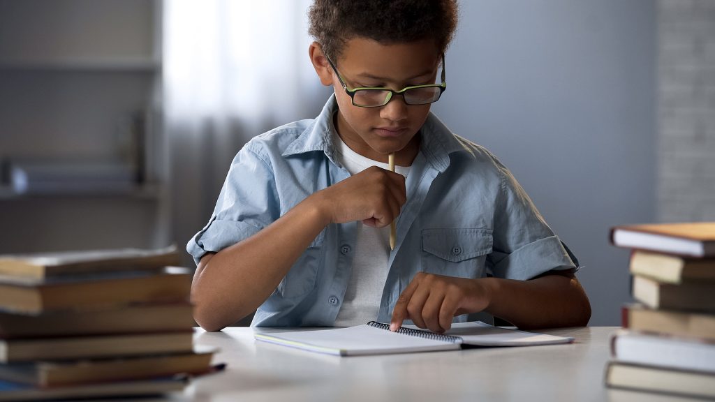 Black boy writes on a piece of paper surrounded by books. He goes to fit homeschool in st. louis, MO at Fit Learning and gets tutoring in St. Louis, MO