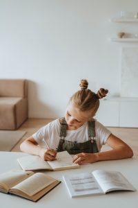young girl wearing overalls sits at a table and writes in her notebook, having benefitted from fluency training at Fit Learning in St. Louis