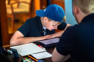 middle-school boy wearing blue cap looks down at his tablet while sitting across from Fit Learning tutor