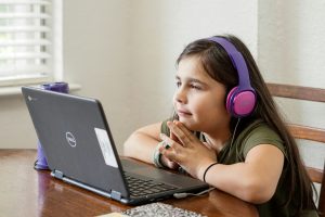 girl wearing purple headphones sits at a table and looks at her laptop during a virtual learning session with her Fit Learning coach