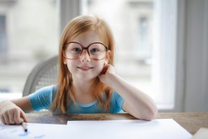 girl with red hair and large glasses looks up from her writing and smiles, proud of her improved reading skills after phonics tutoring at Fit Learning