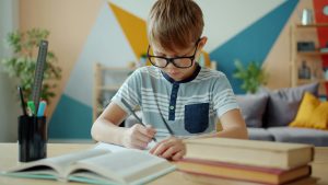 Boy with glasses sits at a desk and writes while looking down at his paper and with a serious expression