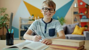 Boy with glasses sits at a desk and looks up from what he's writing with a confident smile