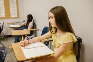 Middle school girl wearing yellow sits in the back of the classroom and looks down at what she's writiing, her facial expression appearing serious and discouraged