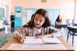 Girl with long dark hair sits at her classroom desk and writes in a notebook, looking engrossed in her work