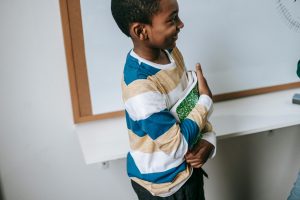 Young boy stands in front of whiteboard holding a green notebook and smiling up at teacher