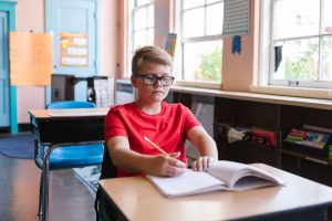 Middle-school boy wearing glasses sits at a desk in his classroom and looks down at his schoolwork with a serious expression
