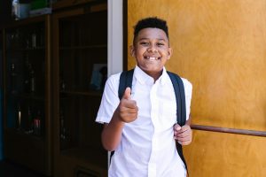 Smiling boy wearing a backpack stands outside school and gives a thumbs-up
