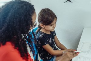 As tutor looks on, young girl sits at desk and looks down at instructional materials