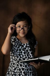 Young girl with dark hair smiles as she holds an open book and looks over her glasses